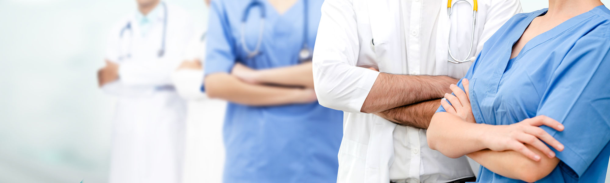 Vier medizinische Fachkräfte in weißen Kitteln und blauen Kasacks stehen mit verschränkten Armen nebeneinander. Die Bildkomposition legt den Fokus auf Selbstbewusstsein und Teamgeist im Gesundheitswesen.